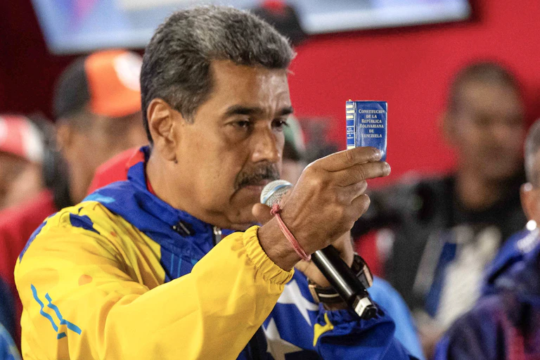 Nicolás Maduro speaking into a microphone while holding a piece of blue paper aloft.