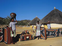 Three people stand in front of houses painted with brown and black designs