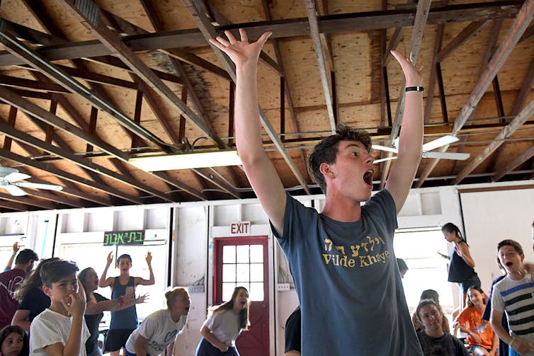 A young man in a blue T-shirt puts his hands in the air as he leads a group of cheering children.