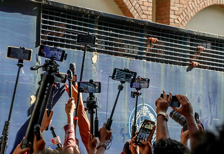 Journalists holding cameras up to a police vehicle transporting prisoners.