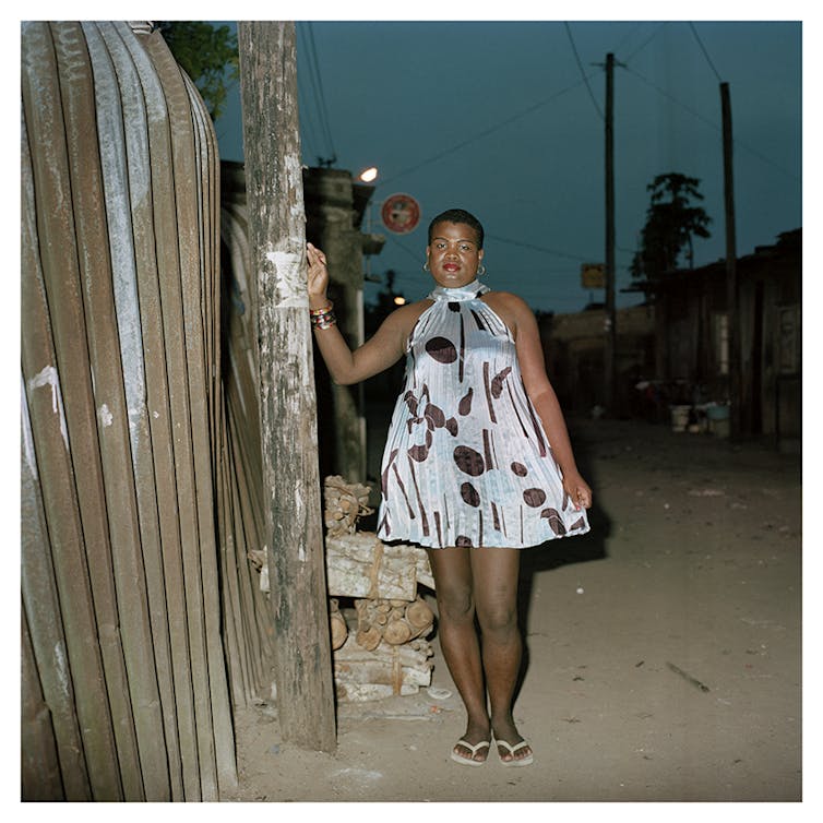 A woman in a short A-line dress poses at night on a dusty road next to a fence.