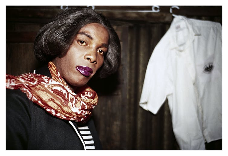 An African woman with a red and white scarf around her neck looks curiously at the camera.