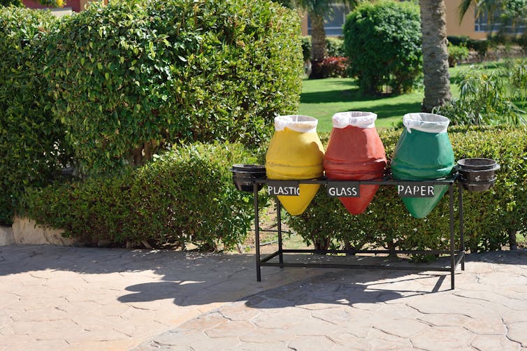 green hedge and grey path, three recycling bins, yellow for plastic, red for glass, green for paper