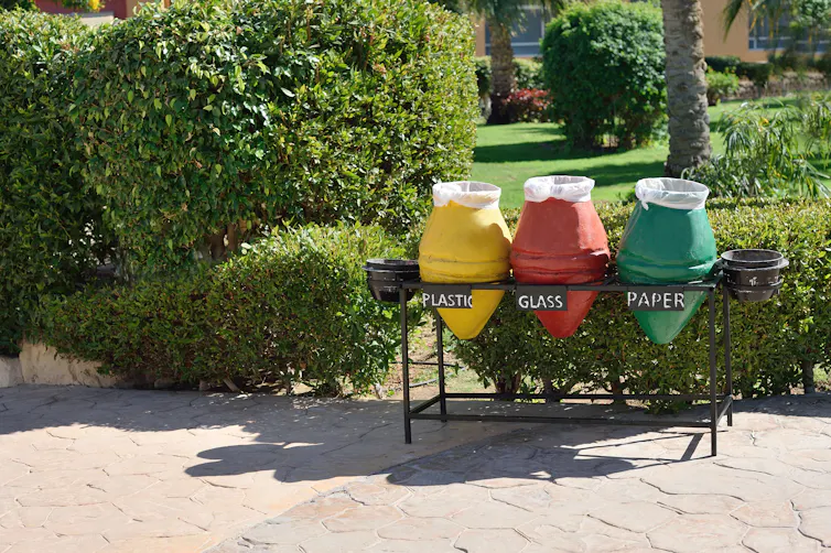 green hedge and grey path, three recycling bins, yellow for plastic, red for glass, green for paper