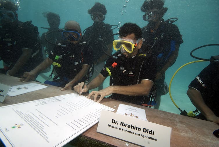 A man in scuba gear sits at a desk underwater and signs government documents in front of him.