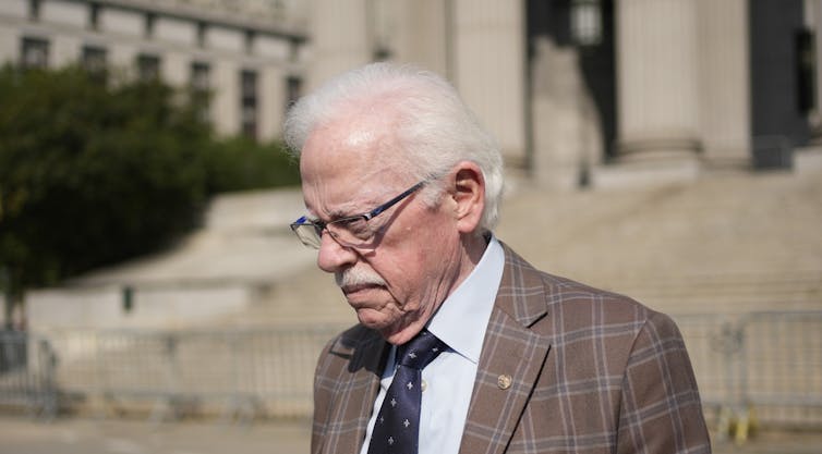 Man with thinning white hair wearing a plaid blazer looks downward while descending an outdoor staircase.