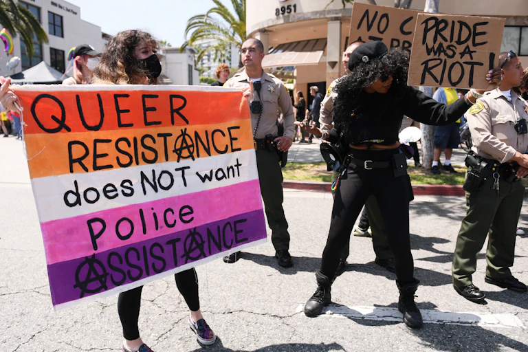 Woman in a face mask holds a sign reading 'queer resistance does not want police assistance' while another woman holds a sign reading 'Pride was a riot.' Uniformed police are standing nearby.
