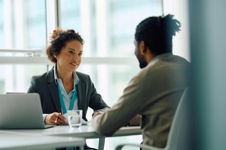A woman in a blazer and lanyard speaks to a man who is seated across from her