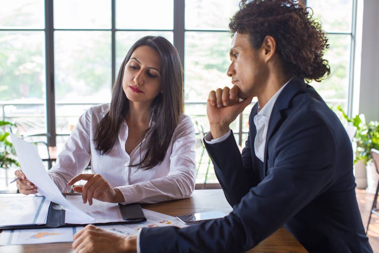 A man and a woman look pensively at a piece of paper being held by the woman. They are both in office attire and are sitting at a desk in an office building.