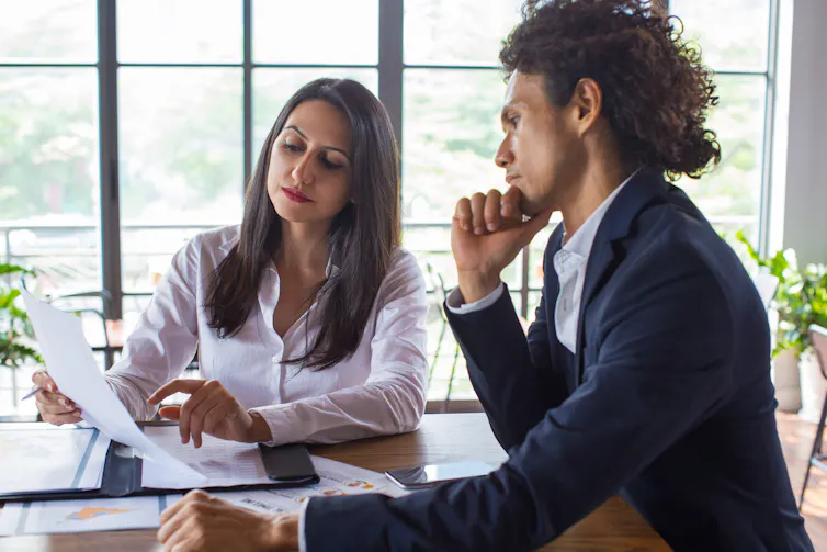A man and a woman look pensively at a piece of paper being held by the woman. They are both in office attire and are sitting at a desk in an office building.
