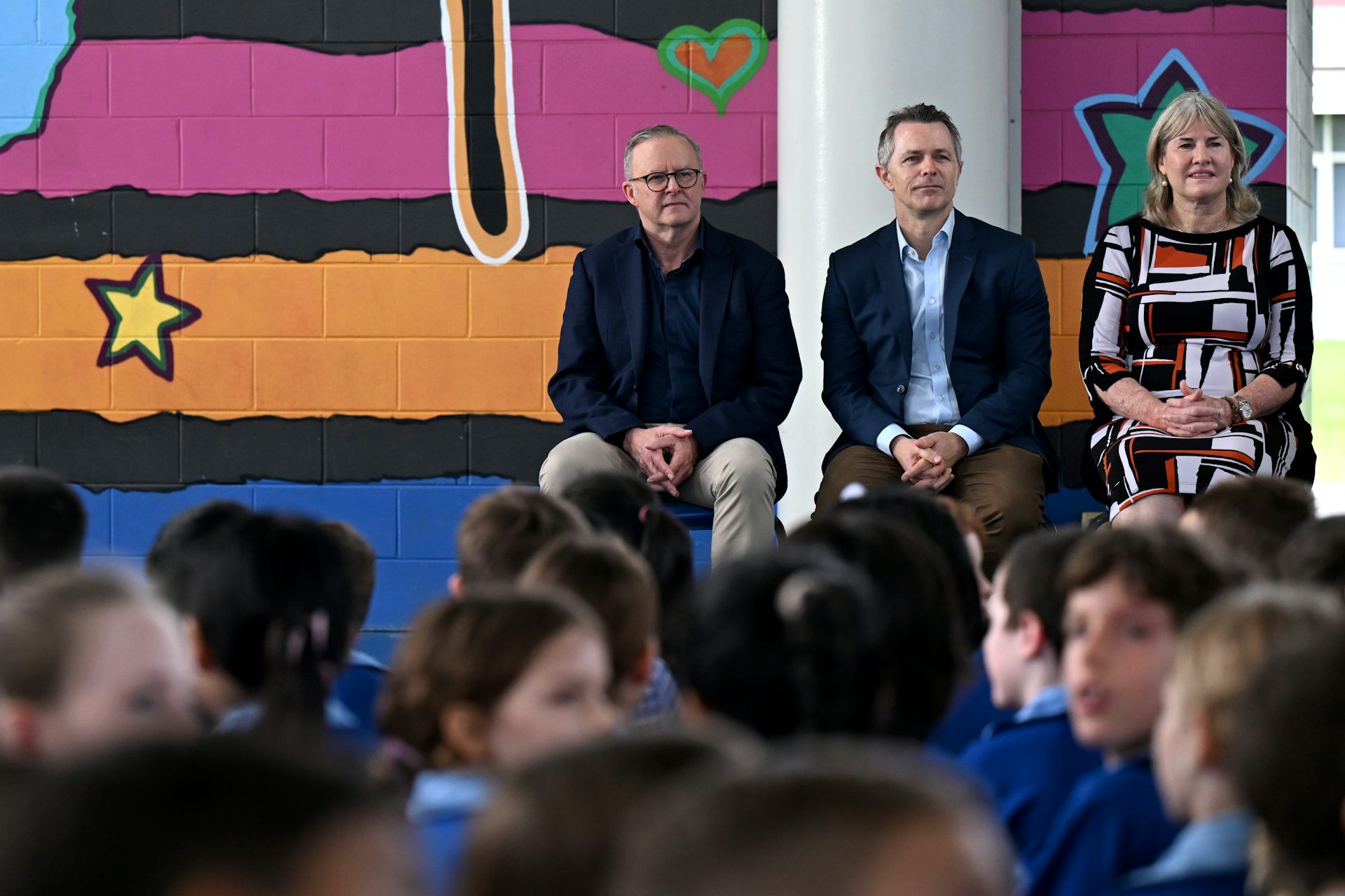 Prime Minister Anthony Albanese, Education Minister Jason Clare and NT Chief Minister Eva Lawler sit on chairs in front of a crowd of primary students, seated on the floor. 