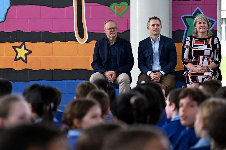 Prime Minister Anthony Albanese, Education Minister Jason Clare and NT Chief Minister Eva Lawler sit on chairs in front of a crowd of primary students, seated on the floor.