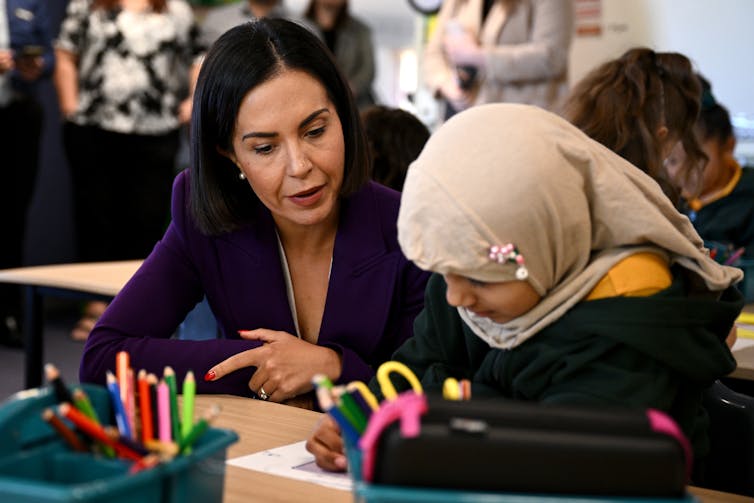 New South Wales Education Minister Prue Car sits at a school table next to a student, who has their head down writing.