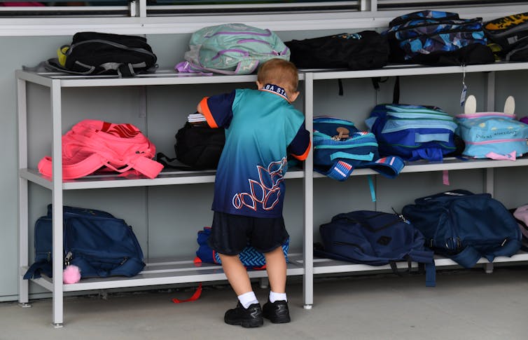 A small child stands in front of a rack of backpacks outside a classroom.