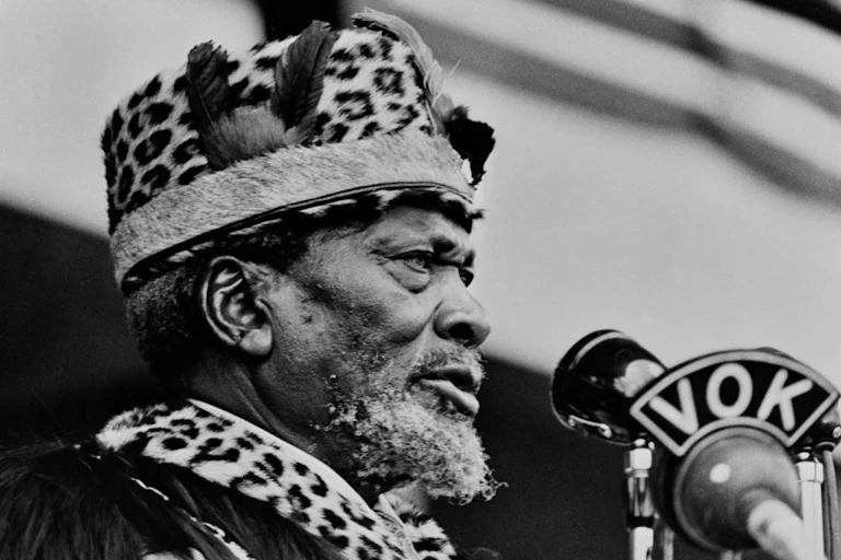A man dressed in a leopard print hat and scarf speaking into a microphone with the letters VOK written on it