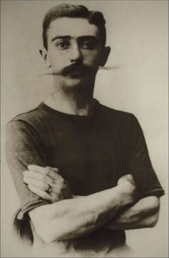 A black and white portrait from the waist up of a man with a long, bushy mustache, posed with his arms crossed over his chest.