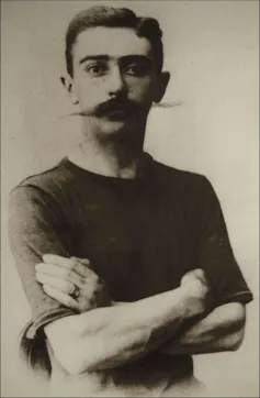 A black and white portrait from the waist up of a man with a long, bushy mustache, posed with his arms crossed over his chest.