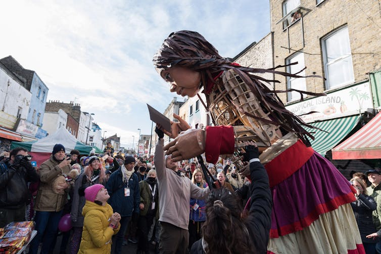 An enormous marionette puppet towers over a small child in the street.