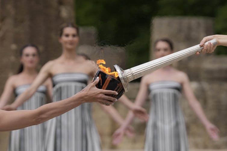 A close-up shot of one person's hand, holding a lighted torch, passing the flame to a dark-colored bowl held by someone else, as three women in striped dresses stand in the background.