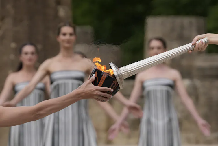 A close-up shot of one person's hand, holding a lighted torch, passing the flame to a dark-colored bowl held by someone else, as three women in striped dresses stand in the background.
