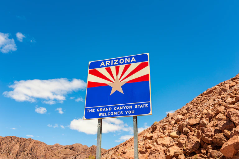 A blue sign with a yellow star and yellow and red stripes says 'Arizona, the Grand Canyon state welcomes you.' A blue sky and red rocks are behind the sign.