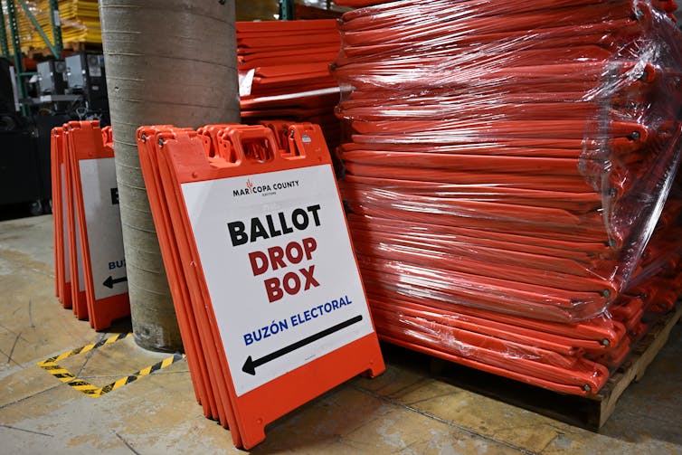 An orange and white sign says, 'Ballot drop box,' and is next to a stack of orange signs that are folded.