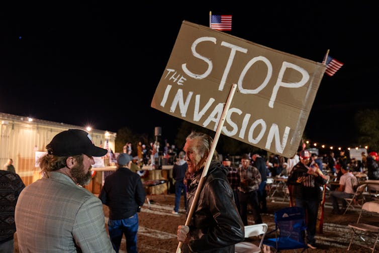 Two men look at each other on a dark night. One holds a sign that says in white paint, 'Stop the invasion.'