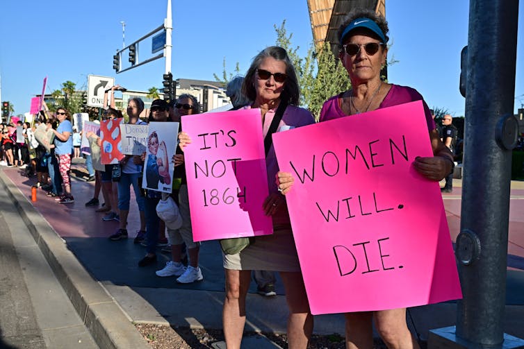 A crowd of people hold signs on a street, including two women who look at the camera and have pink signs that say 'It's not 1864' and 'Women will die.'