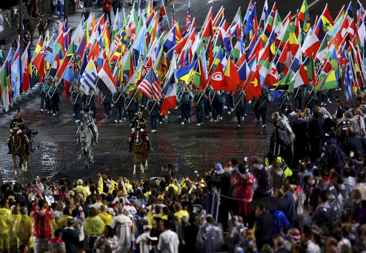 People in uniforms carry dozens of countries' flags as they walk behind three men on horseback, and a crowd watches.