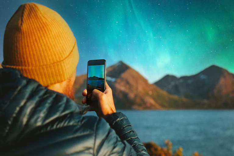 A man in a beanie takes a photo of an aurora and mountain landscape with his smartphone.