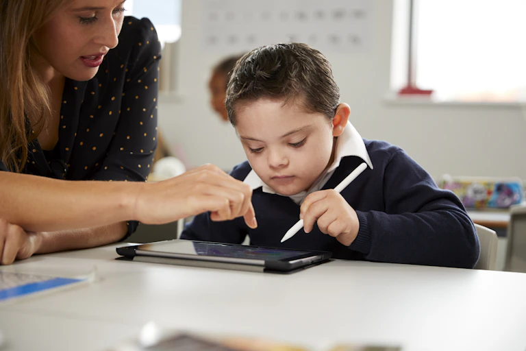 A woman leans over a desk. A young boy uses a stylus to work on a tablet.