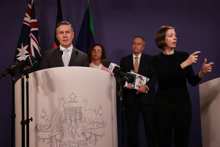 Health Minister Mark Butler speaks at a lectern. A woman standing next to him translating in Auslan. Social Services Minister Amanda Rishwroth and NDIS Minister Bill Shorten stand behind them.