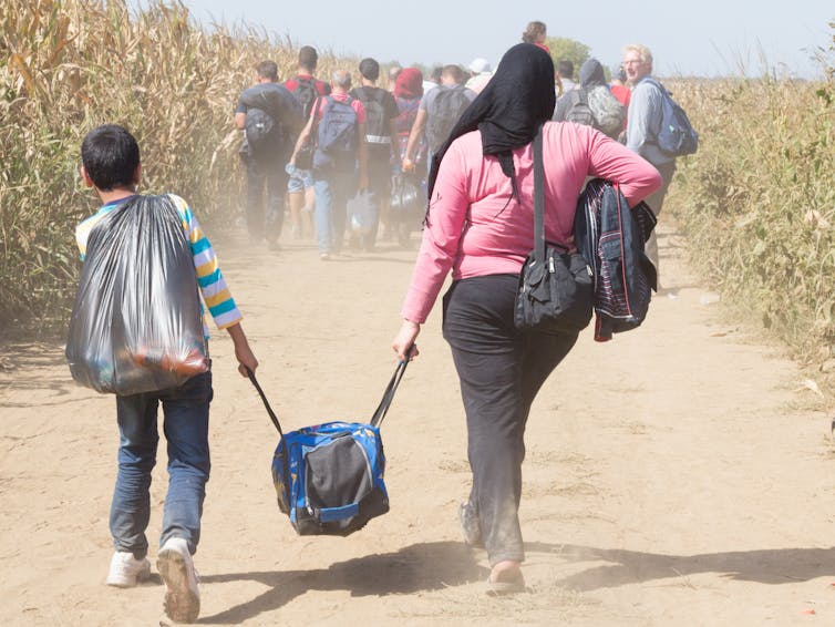 View from behind of a young boy and woman in a head covering walking on a dusty path through a field while carrying a suitcase together