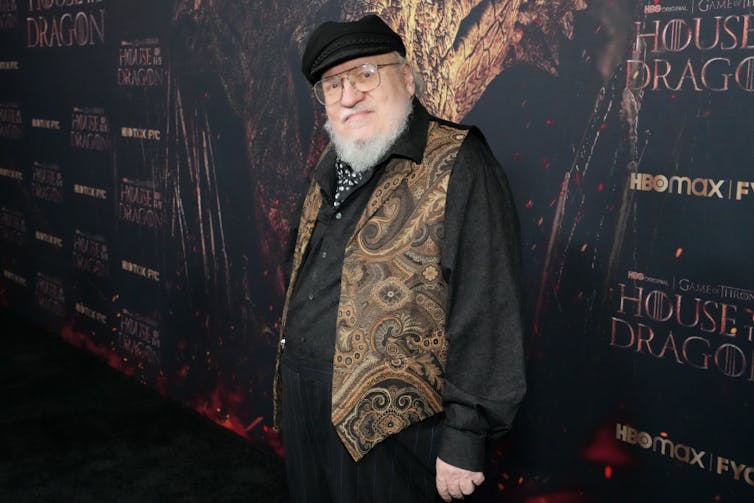 Portly man with white beard, glasses and a black flat cap poses on a red carpet.