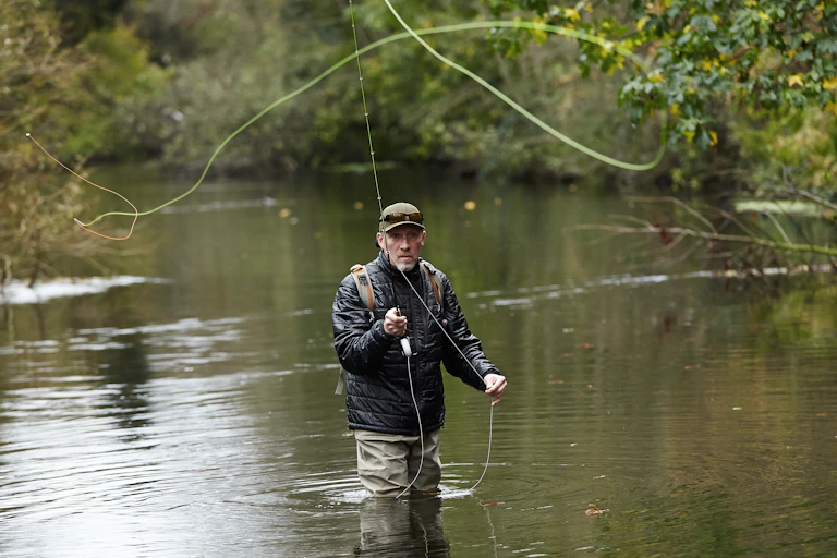man in waders standing in river, fly fishing with fishing rod and line, trees by riverbank