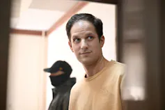 A white man stands near a masked guard behind a glass enclosure.