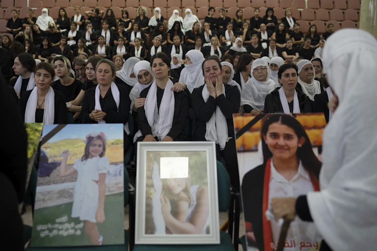 Rows of women in black dresses and white scarves cry as they sit behind large photographs of young girls.
