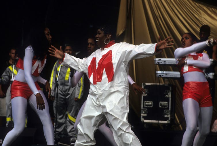 Woman wearing white jumpsuit with large red letter M on chest stands in front of backup dancers onstage