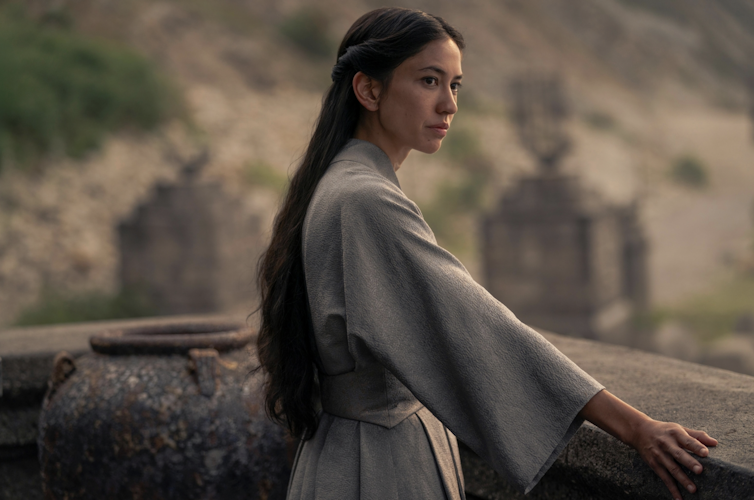 Young woman with long hair stands at a balcony and gazes into the distance.
