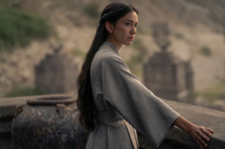 Young woman with long hair stands at a balcony and gazes into the distance.