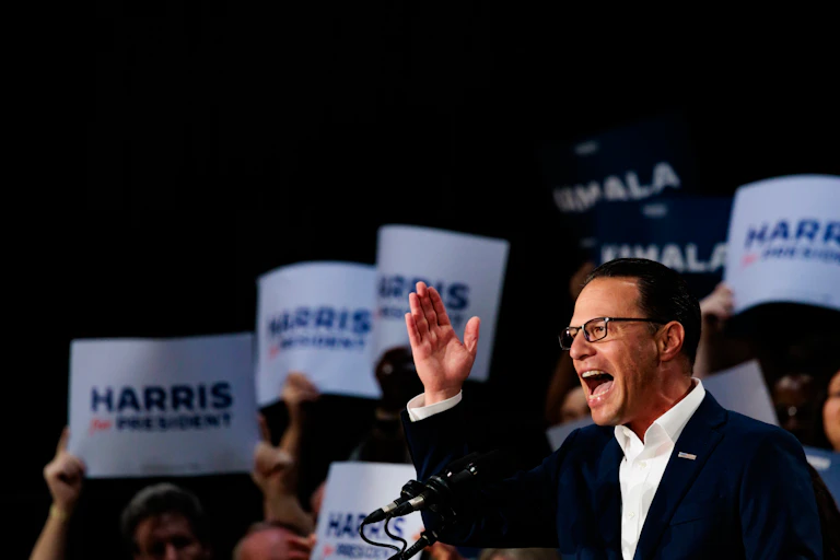 Man wearing suit jacket, dress shirt and glasses speaks at lectern while supporters behind him hold 'Harris for President' signs