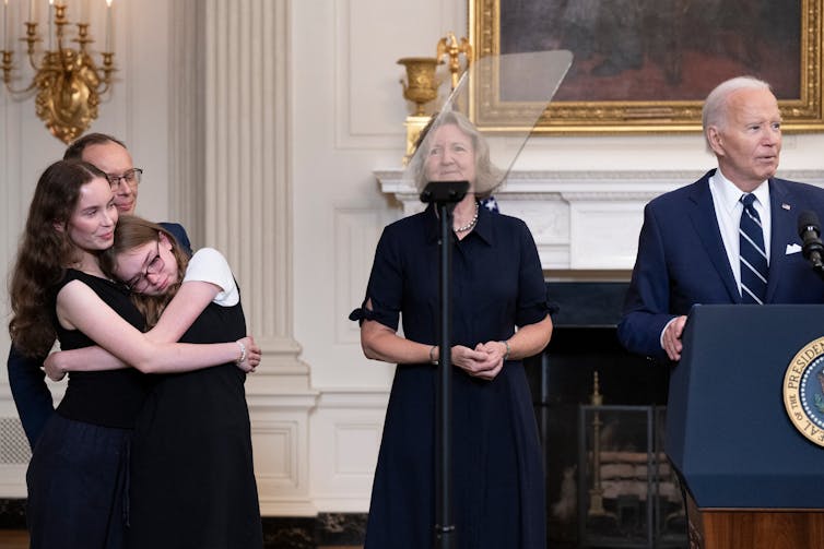 Two young women embrace each other and stand next to two other people in dark clothing, as well as a man with white hair and a blue suit who stands at a podium.