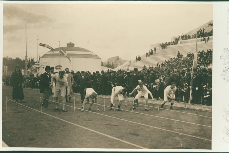 A black and white photo shows five runners in white uniforms crouched in front of lanes marked off with rope, as a crowd watches in stands.