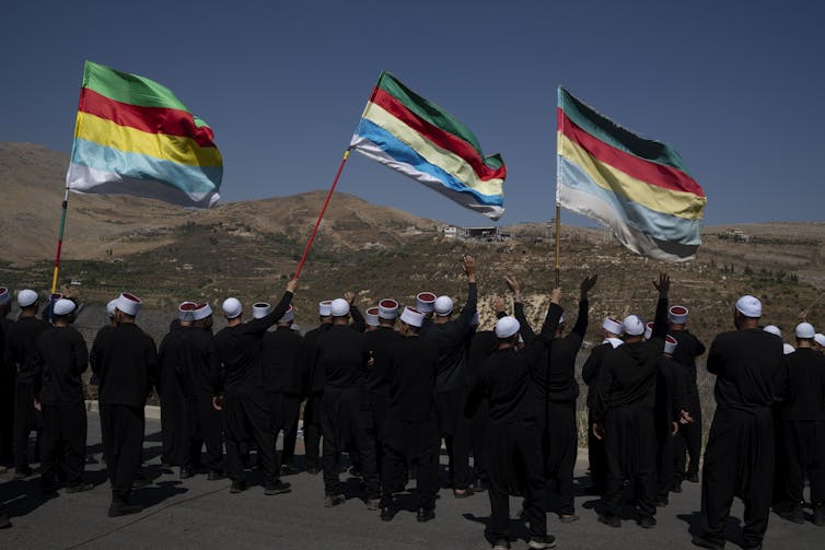 A small crowd of men in black outfits and white hats wave three colorful flags as they look out across a dry, hilly landscape.