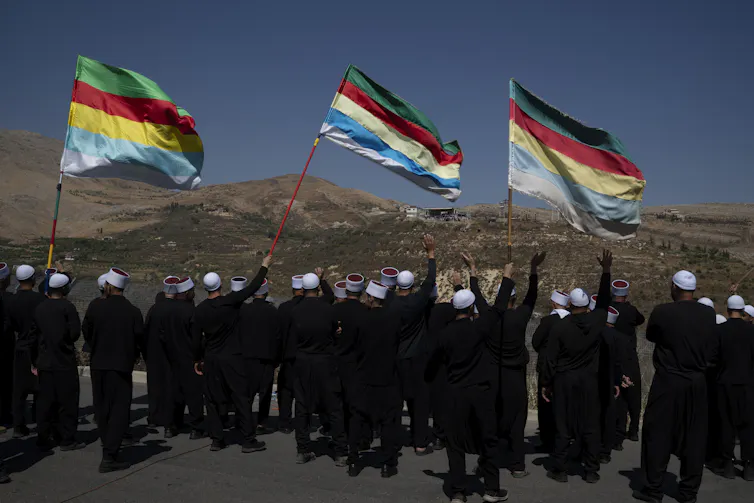 A small crowd of men in black outfits and white hats wave three colorful flags as they look out across a dry, hilly landscape.