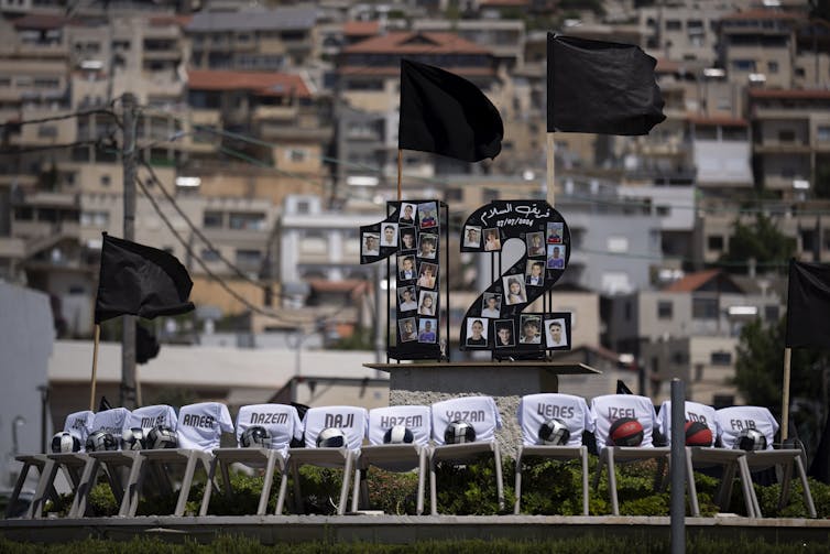 Twelve plastic chairs sit in a row on a street roundabout, covered with white cloths with names written on them.