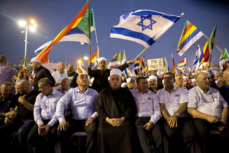 A row of men sit in front of a crowd of people standing and waving flags.
