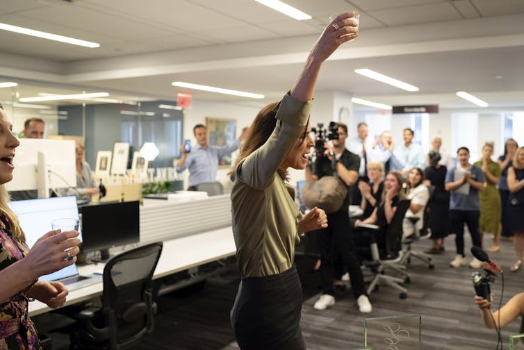 A woman in a newsroom holds a drink high above her head