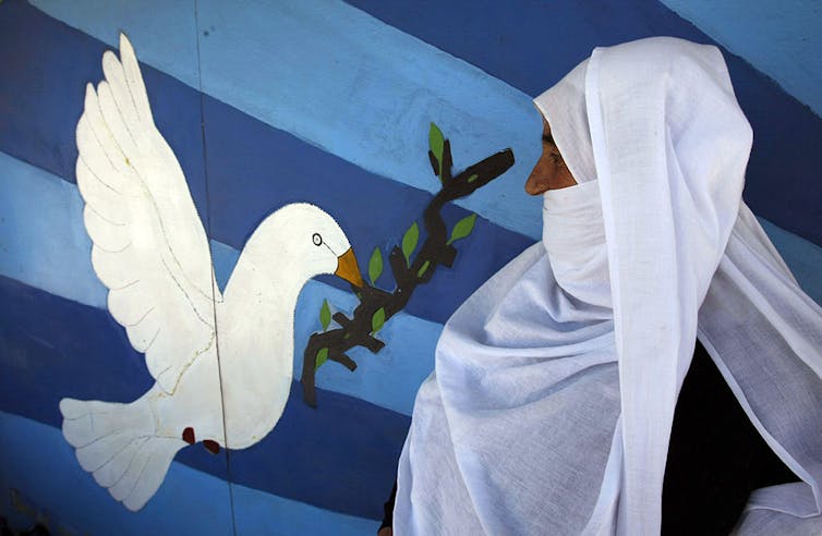 A woman whose head is covered with a large white scarf stands in front of a mural with blue stripes and a picture of a dove holding an olive branch.