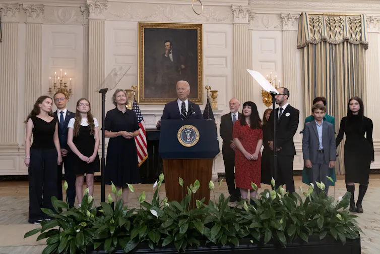A man speaks at a lectern surrounded by people standing next to him